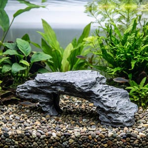 May include: Gray aquarium rock formation with an archway, surrounded by green aquatic plants and small fish. The rock has a textured surface and sits on a bed of small, colorful pebbles.