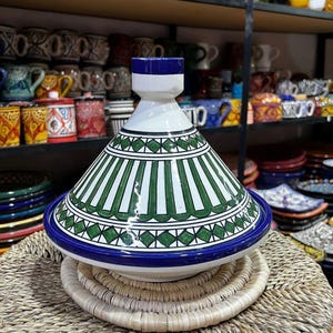 May include: A ceramic tagine pot with a conical lid, featuring a white base with green and white stripes and a blue rim. The pot sits on a woven mat, with shelves of colorful ceramics in the background.