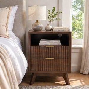 May include: A dark brown wooden nightstand with two drawers and a shelf. A lamp with a beige shade sits on top, along with books and a small vase with greenery. The drawers have a textured, ribbed design and gold-colored handles.