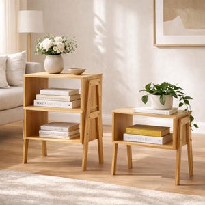 May include: Two light-colored wooden side tables with open shelving. One table has two shelves with books and a vase of white flowers. The other table has one shelf with books and a potted plant. The tables are approximately 24 inches tall.