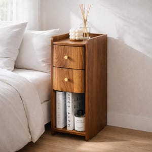 May include: A wooden nightstand with two drawers and a lower shelf. The nightstand is made of a warm-toned wood and features gold-colored knobs. The top of the nightstand holds a candle and a diffuser. The nightstand is next to a bed with white bedding.