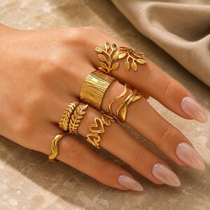 May include: A collection of gold-toned rings worn on fingers. The set includes a wide textured band, a ring with a leaf-like design, a wavy band, a ring featuring open heart shapes, and a simple smooth band, all crafted in a polished metallic finish.