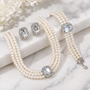 Puede incluir: Un conjunto de joyería que consta de un collar de perlas de tres hilos, una pulsera de perlas de tres hilos y dos pendientes rectangulares. Cada pieza presenta un cristal transparente de corte esmeralda rodeado de pequeños brillantes en metal plateado.
