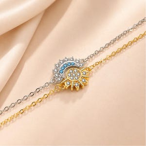 May include: A dual-strand bracelet featuring a silver-toned chain with a crescent moon charm adorned with clear and light blue crystals, and a gold-toned chain with a sun charm featuring clear crystals set in a circular pattern at the center.