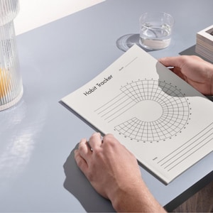 Könnte beinhalten: Ein weißes Notizbuch zur Gewohnheitsverfolgung mit dem Titel "Habit Tracker" wird geöffnet gehalten. Das Notizbuch hat ein kreisförmiges Rasterdesign zur Verfolgung von Gewohnheiten. Ein Glas Wasser und ein dekorativer Glasbehälter sind ebenfalls zu sehen.