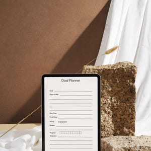 May include: A digital tablet displaying a "Goal Planner" template. The tablet is black with a white screen, featuring sections for goals, steps, dates, and progress. The background includes a brown surface, a textured stone, and white fabric.
