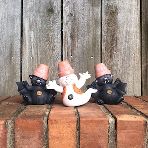 May include: Three Halloween figurines on a brick surface. Two black bat figures with orange pot hats flank a white ghost figure with orange accents. The background is a weathered wooden fence.