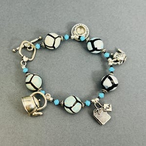 May include: A silver-toned bracelet featuring a toggle clasp, adorned with white and black patterned beads, turquoise accents, and charms. Charms include a teacup, a heart-shaped tag with "TEA" text, and a rabbit.