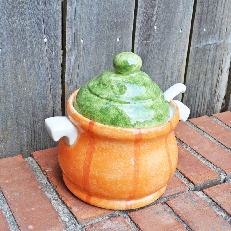 May include: A ceramic soup tureen with a green lid and a small knob. The orange pot has vertical orange stripes and white handles. A white spoon rests on the side. The tureen sits on a brick surface.