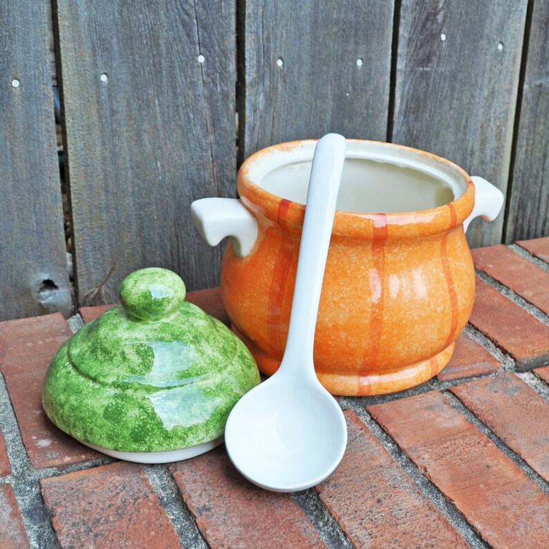 May include: An orange and white ceramic soup tureen with a green lid and a white ladle. The tureen has two handles and vertical orange stripes. The lid is green with a round knob. The ladle is white and rests on a red brick surface.