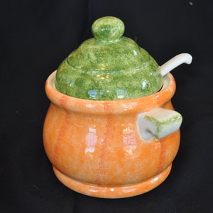 May include: A ceramic sugar bowl with a green lid and a small white spoon. The bowl is orange with vertical red stripes. The lid has a green, mottled finish. A small handle is on the side of the bowl.