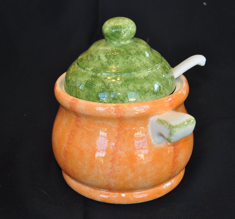 May include: A ceramic sugar bowl with a green lid and a small white spoon. The bowl is orange with vertical red stripes. The lid has a green, mottled finish. A small handle is on the side of the bowl.