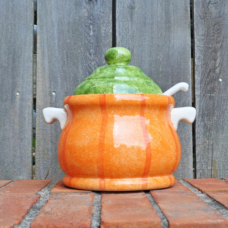 May include: A ceramic soup tureen with a green lid and orange body. The tureen has vertical orange stripes and white handles. A white spoon rests on the rim. The tureen is on a brick surface with a wooden fence in the background.
