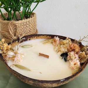 Peut inclure: Une bougie blanche dans un bol en coque de noix de coco brun foncé avec une mèche en bois rectangulaire. La surface de la cire est ornée d'un arrangement de fleurs séchées dans des tons crème, rose pâle et beige, incluant des petites fleurs.