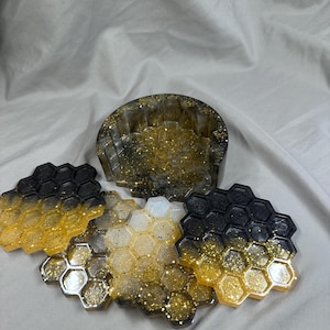 Puede incluir: Un juego de posavasos y un soporte de resina. Los posavasos hexagonales tienen un diseño de panal, con un degradado de resina negra, dorada y transparente, y purpurina dorada. El soporte tiene un diseño similar. Para la decoración del hogar.
