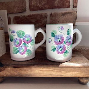 May include: Two white ceramic mugs with floral designs. The mugs feature a pattern of purple and blue flowers with green leaves and stems. The mugs are displayed on a wooden tray with rounded feet.
