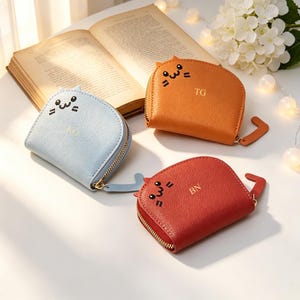 May include: Three cat-shaped coin purses in light blue, orange, and red. Each has a zip closure, a tail-shaped pull, and a cat face design. The purses have initials embroidered on them. A book and flowers are in the background.
