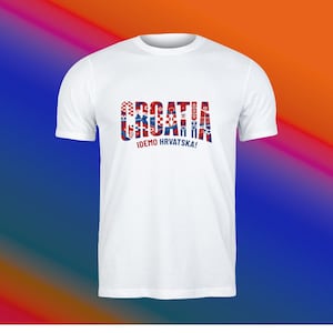 May include: White t-shirt featuring the word "CROATIA" in red, white, and blue, with a Croatian flag design. Below the word is the phrase "IDEMO HRVATSKA!" in black. The shirt has short sleeves.