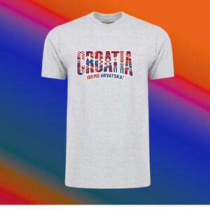 May include: A light gray t-shirt with the word "CROATIA" in a red, white, and blue design across the chest. Below the word is the text "IDEMO HRVATSKA!" in blue. The shirt has short sleeves and a crew neck.