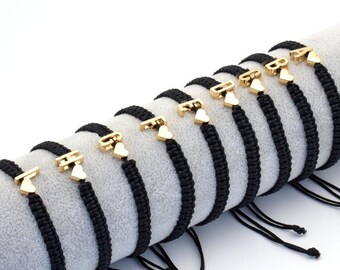 May include: A series of black braided cord bracelets, each featuring a gold-toned metal letter charm paired with a small gold-toned heart charm. The letters displayed on the bracelets are L, H, G, F, E, D, C, B, and A, arranged in a row.