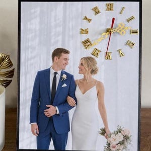 May include: A rectangular wall clock featuring a printed photograph of a person in a blue suit and a person in a white dress holding a bouquet. The clock face has gold-colored Roman numerals, ornate gold hands, and a thin red second hand.