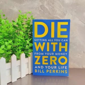 May include: A book titled Die With Zero: Getting All You Can From Your Money And Your Life by Bill Perkins. The cover is blue with large yellow text. The book is positioned next to a small white picket fence planter containing green foliage.