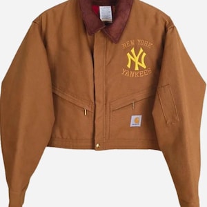 Può includere: Giacca da lavoro marrone con colletto in velluto a coste e logo dei New York Yankees ricamato in giallo. Presenta due tasche frontali, una piccola tasca sul petto e un'etichetta Carhartt. La giacca è appesa a una gruccia di legno.