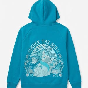 Puede incluir: Sudadera con capucha de color turquesa con un gráfico de una sirena, un pez y el texto "UNDER THE SEA". El diseño es en tonos azules y blancos. La sudadera tiene capucha y mangas largas.