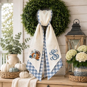 May include: A decorative door hanger featuring a cream-colored fabric sash with a blue and white gingham border. The sash displays an embroidered pumpkin with a football design on one side and a large, navy blue embroidered letter S monogram.