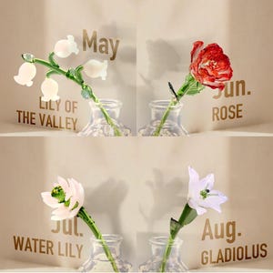 May include: Four glass flowers in clear glass vases, each labeled with a month and name: May Lily of the Valley with white bell-shaped blooms, Jun. Rose with red petals, Jul. Water Lily with pale pink petals, and Aug. Gladiolus with white petals.
