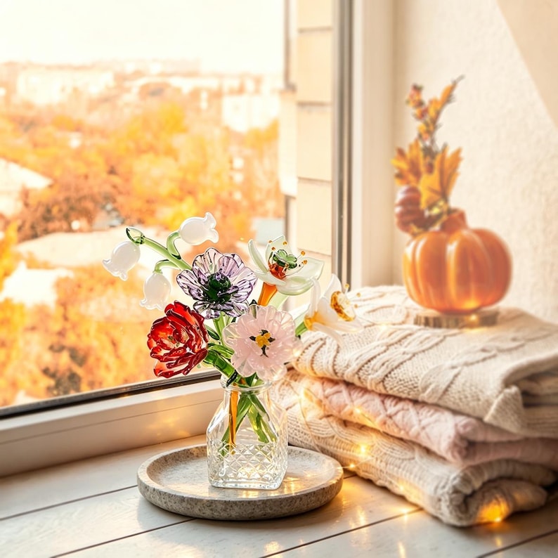 May include: A clear glass vase with a diamond-patterned base holds a bouquet of colorful glass flowers, including white lily of the valley, a purple anemone, a red tulip, a pink blossom, and a white daffodil, all with green glass stems.