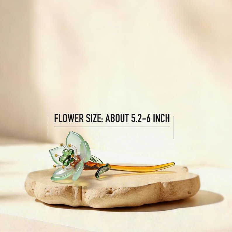 May include: A glass flower ornament with translucent pale green petals, a dark green center, and a long amber-colored stem. The text FLOWER SIZE: ABOUT 5.2-6 INCH is displayed above the object, indicating its length in imperial units.