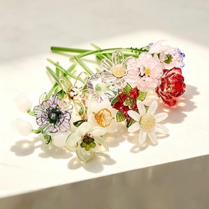 May include: A collection of handcrafted glass flowers featuring various shapes and colors, including red, pink, white, purple, and green petals with clear glass stems. The arrangement includes daisy-like blooms, bell-shaped flowers, and buds.