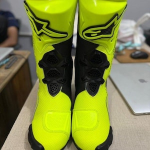 May include: Pair of neon yellow and black motorcycle boots. The boots have a tall, protective design with a textured finish. Black accents and logos are visible on the boots. The boots are designed for safety and performance.