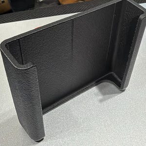 May include: A black, U-shaped plastic holder with a textured, matte finish. The object features a wide, open rectangular interior space with vertical side walls and a flat base, constructed with visible 3D printed layer lines.