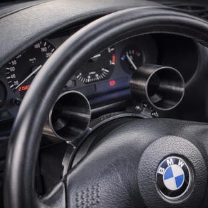 Puede incluir: Primer plano de un volante negro con un soporte de calibre plateado. El soporte del calibre tiene dos aberturas circulares. El salpicadero y los indicadores son visibles en el fondo. El logotipo de BMW está en el volante.