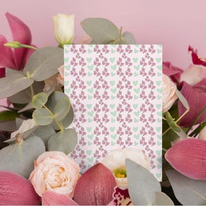 Puede incluir: Una tarjeta rectangular con un patrón de lazos morados y corazones verdes sobre un fondo blanco está rodeada por un ramo de flores rosas y blancas y hojas verdes. El fondo es de color rosa suave.