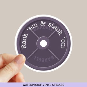 May include: A round, purple barbell weight sticker with the text "Rack 'em & stack 'em" in white. The word "BARBELL" is also printed on the sticker. The sticker is held by a hand. The words "WATERPROOF VINYL STICKER" are printed at the bottom.