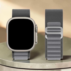 Op de afbeelding: Een zilveren smartwatch met een grijze stoffen band. Het horloge heeft een rechthoekige wijzerplaat met afgeronde hoeken en een zwart scherm. De band is bevestigd met een metalen sluiting.