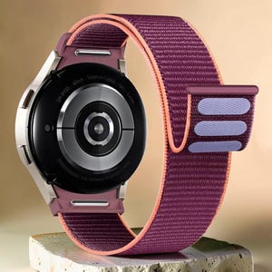 Op de afbeelding: Een smartwatch met een ronde zwarte wijzerplaat en een pruimkleurige stoffen band. De band heeft een perzikkleurige voering en een pruimkleurige sluiting met drie lichtpaarse strepen. De wijzerplaat heeft de tekst "SM-R930 40mm Sapphire Crystal GPS 5ATM".