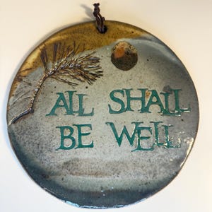 May include: A circular ceramic wall plaque with a mottled grey and brown glaze. It features an embossed pine branch, a small circular indentation, and the green text "ALL SHALL BE WELL" in the center. A dark brown leather cord is attached at the top.