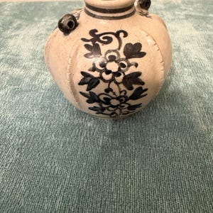 May include: A small, round, off-white ceramic vase featuring a hand-painted dark blue floral pattern. The vessel has a narrow neck with a dark blue rim, a bulbous body with vertical ridges, and two small, dark blue circular handles on the shoulders.