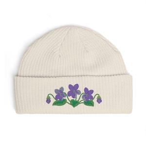 May include: A cream-colored ribbed knit beanie featuring an embroidered design of purple violet flowers with green leaves and stems centered on the folded cuff. The hat is constructed from a thick, textured yarn with a classic cuffed brim.