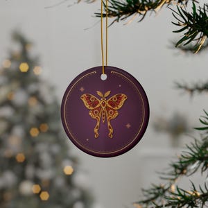 May include: A round, deep purple ceramic ornament featuring a gold and dark red moth illustration at the center. The moth design includes intricate filigree patterns and small circular accents. The ornament hangs from a thin gold string.