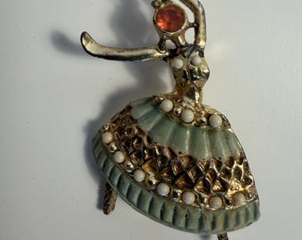 May include: A gold-toned metal brooch shaped like a dancing figure. The figure features a pale green pleated skirt with a textured gold lattice pattern, accented by small white beads. An orange circular stone sits at the head.