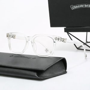 Peut inclure: Lunettes en acétate transparent avec des emblèmes en forme de croix argentée sur les branches et les charnières. La monture carrée est présentée avec un étui en cuir noir et une boîte noire portant le logo Chrome Hearts imprimé en blanc.