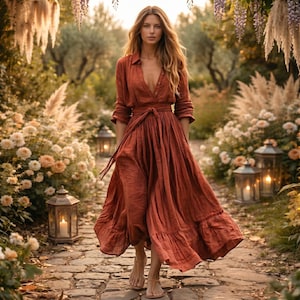 Puede incluir: Un vestido largo de color óxido con una parte superior abotonada y una falda fluida. El vestido presenta una cintura anudada y mangas largas. La mujer camina por un camino de piedra en un jardín, rodeada de flores y faroles.