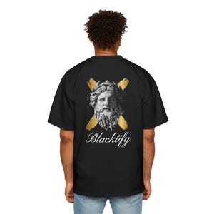 Puede incluir: Camiseta negra con un busto blanco de un hombre barbudo con una corona de laurel, sobre dos cruces doradas. La palabra "Blacktify" está escrita en cursiva debajo del busto.