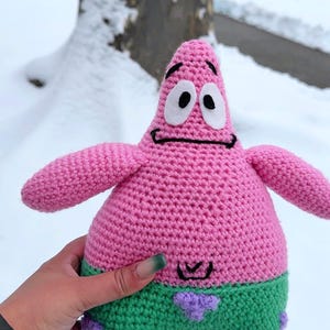 May include: A handmade crochet Patrick Star plush toy. The toy is pink with green shorts and purple accents. It has black eyes and a black mouth. The background is a snowy outdoor scene. The toy is a character from Spongebob Squarepants.