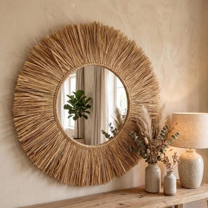 May include: A round mirror with a natural woven straw frame. The mirror is surrounded by a sunburst design, creating a rustic and bohemian aesthetic. The mirror is displayed above a wooden console table, with decorative vases and a lamp.